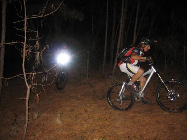 Two mountain bikers navigate a dark, wooded trail at night. One rider, wearing a helmet and bright clothing, is in focus, while the second rider is visible in the background, illuminated by a bright light. The forest floor is covered with pine needles, and trees surround the trail. Blue Clay Bike Park mountain bike trail.