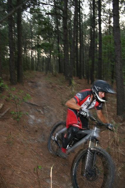 A mountain biker in a red and black outfit rides through a wooded trail, maneuvering around trees and brush. The sunlight filters through the leaves, casting a dappled light on the path as the cyclist leans into the turn. Blue Clay Bike Park mountain bike trail.