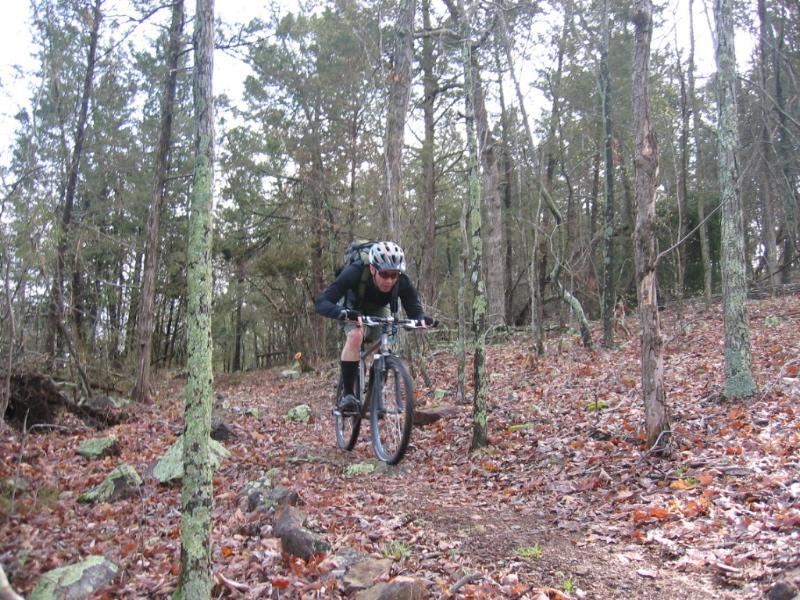 A mountain biker rides swiftly through a forest trail covered in fallen leaves and surrounded by trees. The rider, dressed in a black outfit and helmet, is positioned low on the bike, showcasing an active and dynamic posture. Council Bluffs Trail mountain bike trail.