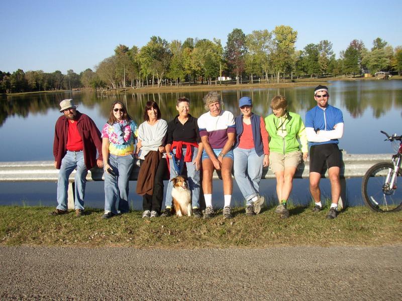 A group of eight people stands by a lake, enjoying a sunny day. They are casually dressed and smiling, with a dog sitting in front of one person. The landscape features trees in the background and a calm water surface reflecting the clear sky. A bicycle is parked nearby, and the atmosphere appears relaxed and friendly. Herb Parsons mountain bike trail.