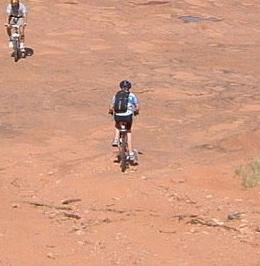 Two cyclists riding on a rocky dirt path in a natural outdoor setting. One cyclist is positioned in the foreground, wearing a blue shirt and a backpack, while the other cyclist is further back on the same trail. The landscape features earthy tones and sparse vegetation. Gemini Bridges mountain bike trail.