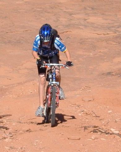 A person riding a mountain bike uphill on a sandy, rocky surface, wearing a blue helmet and a blue cycling jersey, with shorts and cycling shoes. Gemini Bridges mountain bike trail.