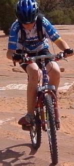 A child wearing a blue helmet and a cycling jersey rides a mountain bike on a rocky trail. The cyclist leans forward as they navigate the terrain, showcasing an active outdoor adventure. Gemini Bridges mountain bike trail.