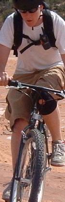 A person riding a mountain bike on a dirt path, wearing sunglasses, a white t-shirt, and shorts. The individual has a backpack and appears focused on biking. Gemini Bridges mountain bike trail.