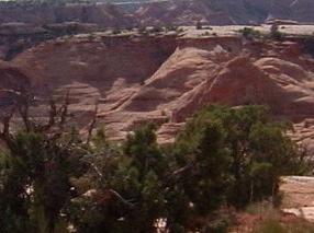 A rocky landscape featuring layered red and brown hills, with sparse vegetation in the foreground and blue sky in the background. Gemini Bridges mountain bike trail.