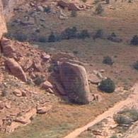 A landscape featuring a large boulder in a rocky, grassy area, with additional smaller rocks and sparse vegetation surrounding it. A dirt path runs along the lower part of the image. Gemini Bridges mountain bike trail.