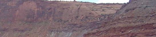 A panoramic view of a rugged red rock cliff, showcasing layered geological formations under a blue sky with scattered clouds. Green vegetation is visible at the cliff's summit, contrasting with the earthy tones of the rock. Gemini Bridges mountain bike trail.