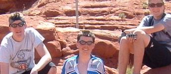 Three young people sitting on red rock formations under sunlight, wearing sunglasses and casual clothing. They appear relaxed and are enjoying a day outdoors. Hurrah Pass mountain bike trail.