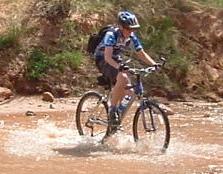 A person riding a mountain bike through shallow water, creating splashes, with rocky terrain and greenery in the background. Hurrah Pass mountain bike trail.