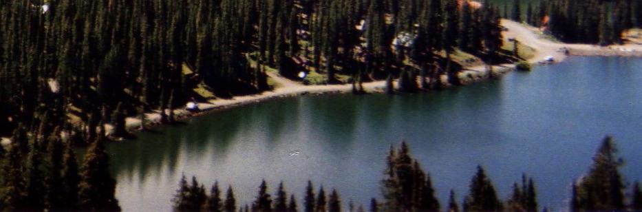 Aerial view of a serene lake surrounded by tall evergreen trees, with a sandy shore and a winding pathway. The water reflects the greenery and the blue sky, creating a peaceful natural scene. Lower Crags Crest Trail mountain bike trail.