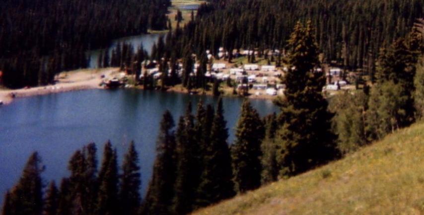 A scenic view of a tranquil lake surrounded by dense evergreen trees, with a cluster of camping trailers visible near the water's edge. The backdrop features a lush green hillside, enhancing the natural beauty of the landscape. Lower Crags Crest Trail mountain bike trail.