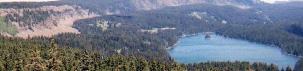 A scenic view of a tranquil lake surrounded by dense forests and distant hills under a clear sky, showcasing the beauty of nature. Lower Crags Crest Trail mountain bike trail.