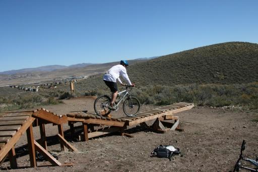 A cyclist in a white shirt and blue helmet is riding a mountain bike over a wooden ramp on a dirt path, with hills and a valley visible in the background. Trailside Loop And Skills Park mountain bike trail.