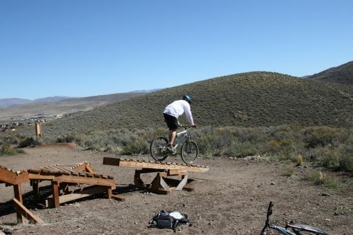 A cyclist in a white shirt and helmet performing a jump on a mountain bike over a wooden ramp, with a hilly landscape in the background under a clear blue sky. Trailside Loop And Skills Park mountain bike trail.