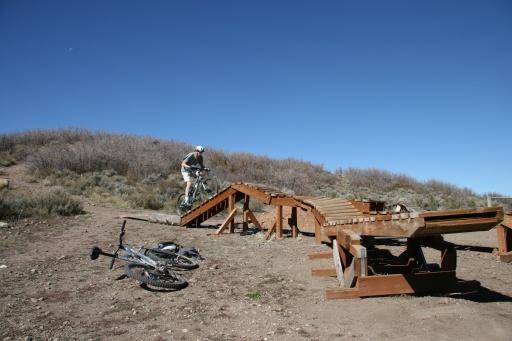 A person rides a bike while jumping off a wooden ramp on a dirt trail, with two bicycles lying on the ground nearby. The background features a hilly landscape and a clear blue sky. Trailside Loop And Skills Park mountain bike trail.