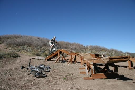 A person riding a mountain bike is jumping off a wooden ramp in a dirt area surrounded by shrubs and plants. Two bicycles are resting on the ground nearby, with a clear blue sky above. Trailside Loop And Skills Park mountain bike trail.