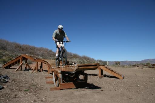 A cyclist performing a trick on a bike at a dirt bike park, navigating a wooden ramp under a clear blue sky. Trailside Loop And Skills Park mountain bike trail.