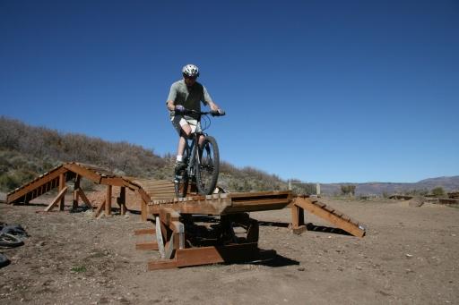 A person riding a mountain bike on a wooden ramp structure in a dirt park, with a clear blue sky in the background. The rider is balancing on the ramp, showcasing a challenging biking maneuver. Trailside Loop And Skills Park mountain bike trail.