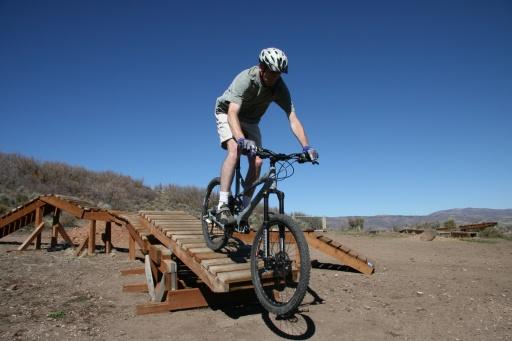 A person riding a mountain bike over a wooden ramp in a dirt area, with clear blue skies and hills in the background. The cyclist is wearing a helmet and athletic clothing. Trailside Loop And Skills Park mountain bike trail.