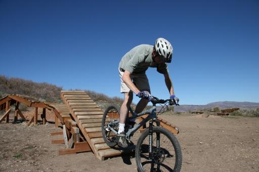 A cyclist wearing a helmet navigates a wooden ramp on a mountain bike, with clear blue skies and a hilly landscape in the background. Trailside Loop And Skills Park mountain bike trail.