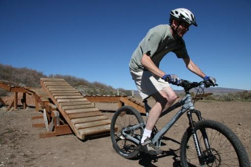 A person riding a mountain bike approaches a wooden ramp on a dirt trail, wearing a helmet and cycling gear, with blue sky and shrubbery in the background. Trailside Loop And Skills Park mountain bike trail.