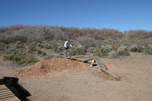 A mountain biker riding over a wooden ramp, preparing to leap off a dirt mound in an outdoor area with sparse vegetation and a clear blue sky. Trailside Loop And Skills Park mountain bike trail.