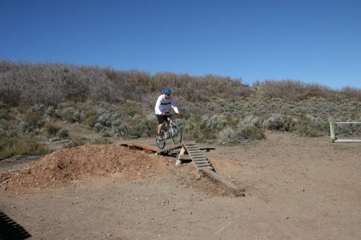 A cyclist in a white long-sleeve shirt and blue helmet is airborne, jumping off a wooden ramp made from planks, over a dirt mound. The background features scrubby vegetation and a clear blue sky. Trailside Loop And Skills Park mountain bike trail.
