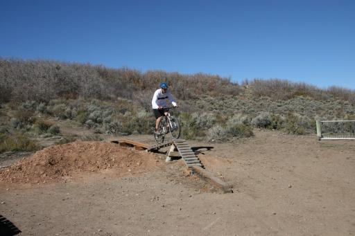 A mountain biker in a blue helmet rides off a wooden ramp onto a dirt path, surrounded by shrubs and a clear blue sky. Trailside Loop And Skills Park mountain bike trail.
