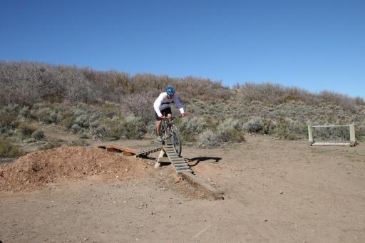 A cyclist performing a jump on a wooden ramp in a dirt area surrounded by shrubs and low vegetation under a clear blue sky. Trailside Loop And Skills Park mountain bike trail.