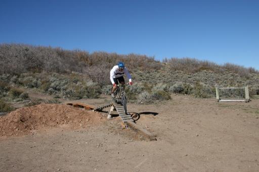 A mountain biker jumps off a wooden ramp, soaring over a dirt mound in a mountainous terrain. The background features shrubs and a clear blue sky. Trailside Loop And Skills Park mountain bike trail.