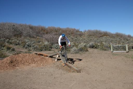 A mountain biker in a white long-sleeve shirt and blue helmet is airborne above a wooden ramp, with a dirt jump in the background. The landscape features sparse vegetation and clear blue skies. Trailside Loop And Skills Park mountain bike trail.