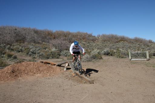 A person riding a mountain bike is skillfully navigating a wooden ramp in a dirt area surrounded by sparse vegetation. In the background, there are low shrubs and a clear blue sky. Trailside Loop And Skills Park mountain bike trail.