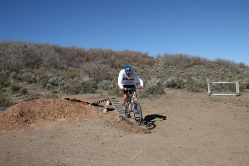 A cyclist in a white long-sleeve shirt and blue helmet is performing a jump on a mountain bike off a wooden ramp, with a dirt and scrubland background under a clear blue sky. Trailside Loop And Skills Park mountain bike trail.