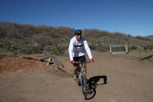 A cyclist riding a mountain bike on a dirt trail, wearing a white long-sleeve shirt and blue helmet. In the background, there are shrubs and a wooden ramp, with a goalpost visible in the distance under a clear blue sky. Trailside Loop And Skills Park mountain bike trail.