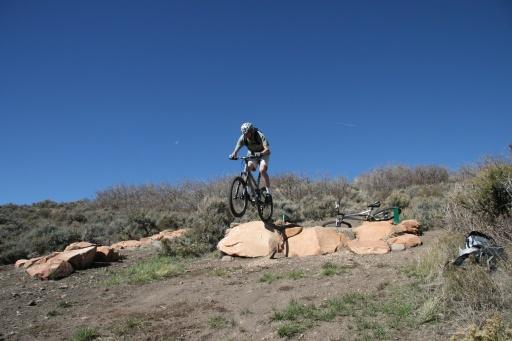 A mountain biker jumping off a rocky outcrop against a clear blue sky, with vegetation and another bike in the background. Trailside Loop And Skills Park mountain bike trail.