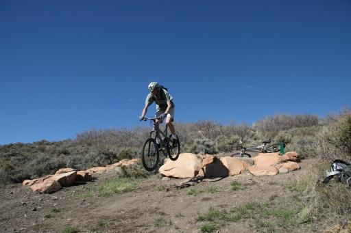A mountain biker performing a jump over a rocky pathway on a sunny day, with blue skies and shrubs in the background. Trailside Loop And Skills Park mountain bike trail.