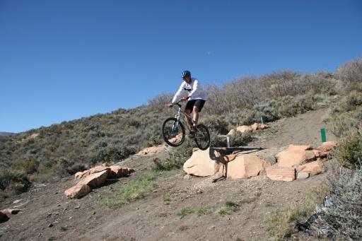 A mountain biker performing a jump on a rocky trail, surrounded by shrubbery and open blue sky. Trailside Loop And Skills Park mountain bike trail.
