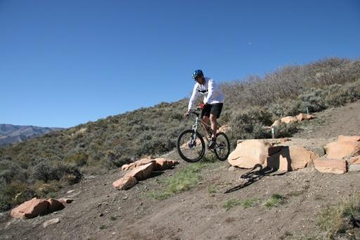 A mountain biker in a white long-sleeve shirt and helmet is jumping off a small rock formation on a dirt trail surrounded by shrubs and hills under a clear blue sky. Trailside Loop And Skills Park mountain bike trail.