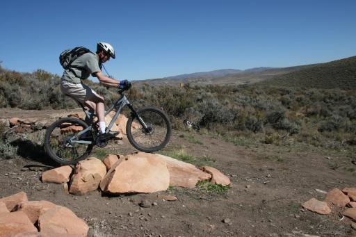 A mountain biker in a helmet and backpack jumps over a rocky obstacle on a dirt trail, surrounded by sparse vegetation and a clear blue sky. The landscape features hills in the background. Trailside Loop And Skills Park mountain bike trail.