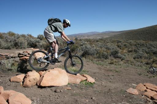 A person in a helmet riding a mountain bike, performing a jump over a large rock while on a dirt trail surrounded by shrubs and hills under a clear blue sky. Trailside Loop And Skills Park mountain bike trail.