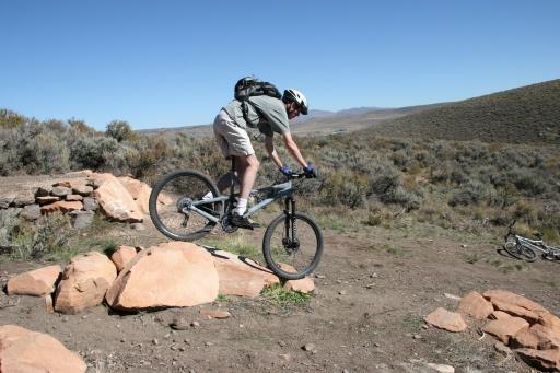 A person in athletic clothing riding a mountain bike over a rocky surface in an outdoor setting, with hills and shrubs in the background. The rider is maneuvering to balance on the bike while navigating the terrain. Trailside Loop And Skills Park mountain bike trail.