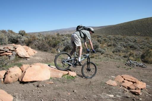A mountain biker performing a trick on rocky terrain, with a hilly landscape in the background under a clear blue sky. Trailside Loop And Skills Park mountain bike trail.