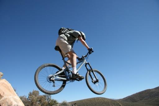 A person performing a jump on a mountain bike over a rocky surface, with a clear blue sky in the background. Trailside Loop And Skills Park mountain bike trail.