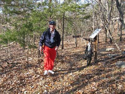 A person wearing a helmet and a jacket is walking near a mountain bike parked next to a sign in a wooded area, with fallen leaves covering the ground. Sunlight filters through the trees, creating a natural environment for outdoor activities. Lost Bridge Trail mountain bike trail.