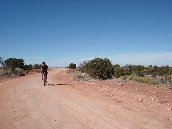 A person riding a bicycle along a dirt road surrounded by sparse vegetation and blue sky. The landscape features reddish soil and scattered shrubs on either side of the path. Gemini Bridges mountain bike trail.