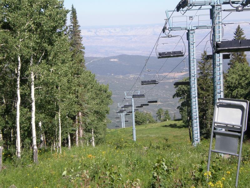 A scenic view of a ski lift ascending a mountain, surrounded by green foliage and trees. In the background, rolling hills and valleys can be seen under a clear sky. The image captures the tranquility of a mountain landscape during summer. West Bench Trail mountain bike trail.