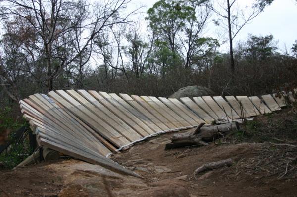 A wooden structure partially collapsed on a dirt path surrounded by sparse trees and underbrush, with some rocks visible in the background. The boards are arranged in a diagonal pattern, creating an angled surface leading down the slope. You Yangs Regional Park mountain bike trail.