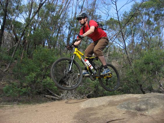 A person in a red shirt and a helmet jumps on a yellow mountain bike over a rocky terrain, surrounded by trees and greenery. You Yangs Regional Park mountain bike trail.