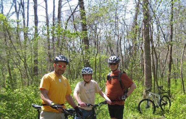 Three individuals wearing helmets and cycling gear are standing on a trail surrounded by trees and greenery. They are smiling and posing with their bicycles, enjoying a sunny day outdoors. The scene captures a moment of recreational biking in nature. Dutton Hollow Loop mountain bike trail.
