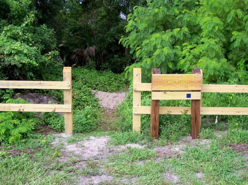 Entrance to a nature sanctuary featuring a wooden fence and a sign that reads "Eagle's Creek Sanctuary - Mountain Bike Trail." Lush greenery surrounds the area, indicating a natural, wooded environment. Turkey Creek mountain bike trail.